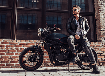 Man wearing a leather jacket and sunglasses leans against his motorcycle that is parked in front of the window of a brick building